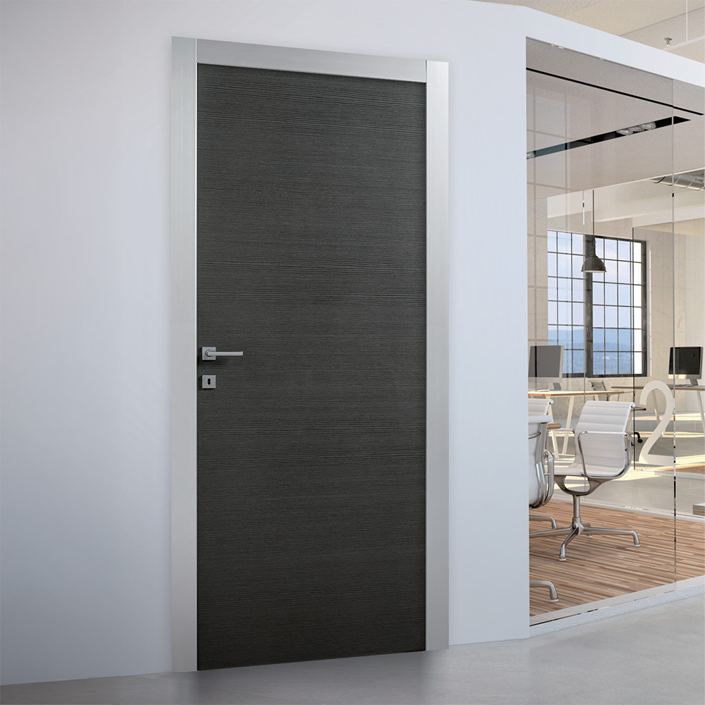 Questa immagine mostra una porta moderna e minimalista, con finitura in legno scuro a venature orizzontali e cornice in metallo satinato. Si trova in un ambiente lavorativo elegante e luminoso, con pareti bianche e pareti divisorie in vetro che lasciano intravedere una sala riunioni con arredi contemporanei.